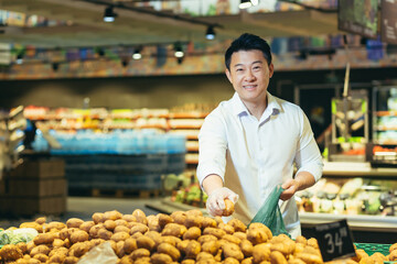 Wall Mural - Portrait of an Asian shopper in the grocery department of a supermarket, a man chooses vegetables and buys potatoes, smiling and looking at the camera