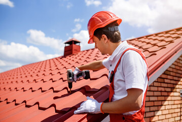 Wall Mural - Man worker master in red overall and helmet is fixing the metal tile roof.