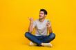© deagreez - Full size photo of impressed brunet young guy sit hold telephone look promo wear t-shirt jeans shoes isolated on yellow background