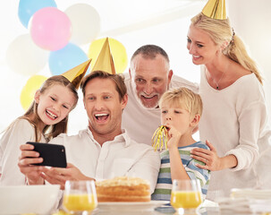 A happy family celebrating a birthday with a party, wearing hats and taking a selfie using a phone. Mature man taking a photo of his father, wife and children at a party while making special memories