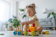 Girl plays with a wooden railway on the living room floor, builds in light interior. Happy cute little baby sits on a light background at home and plays with a wooden zero waste toys