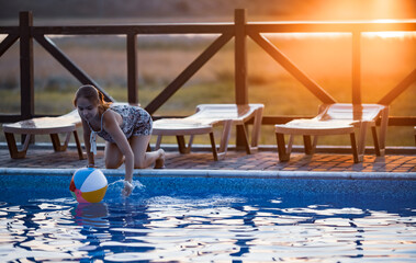 Canvas Print - A girl with hair braided in a bun in bright suit plays by the pool with a ball against the background of summer sun