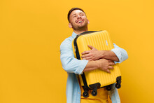 Enjoyed Funny Smiling Tanned Handsome Man In Blue Shirt Hugs Hold Suitcase Ready For Vacation Posing Isolated On Orange Yellow Studio Background. Copy Space Banner Mockup. Trip Journeys Concept