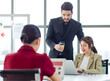 © Bangkok Click Studio - Millennial Indian bearded male businessman mentor standing holding disposable coffee cup teaching coaching Asian intern trainee secretary working with laptop computer in multinational company office