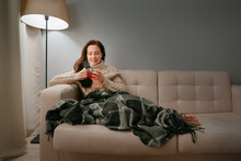 Young woman wrapped herself in a plaid while sitting on a beige sofa drinks hot tea for colds.