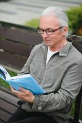 Wall Mural - Old man with gray hair reads a book on a bench in the park. Rest in the park.