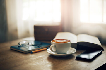 Close up view, Latte coffee in white cup and smart phone on wooden table near bright window. blurred background with book, eyeglasses on blue note book and pen, vintage color tone