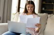 © fizkes - Happy pretty woman reading papers, letter, check documents or domestic bills smile feels satisfied, use laptop, doing paperwork or studying sit on sofa at home. Correspondence, get good news concept