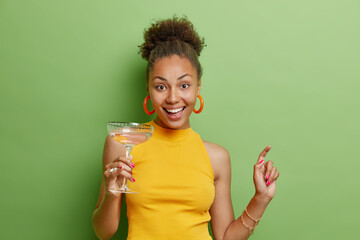 Positive carefree beautiful woman with curly dark hair dances with glass of cocktail wears yellow t shirt and earrings has positive mood isolated over vivid green background enjoys party time