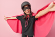 Cheerful woman in superhero costume keeps arms outstretched pretends having super power brave to help someone smiles positively poses against pink background. Mysterious character on masquerade.