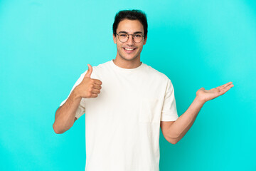 Wall Mural - Caucasian handsome man over isolated blue background holding copyspace imaginary on the palm to insert an ad and with thumbs up