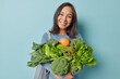 © wayhome.studio  - Positive Asian woman with dark hair carries freshly picked vegetables and fruits net bag on shoulder keeps to healthy diet being vegetarian dressed in sportswear isolated over blue background
