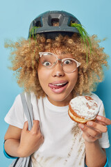 Wall Mural - Vertical shot of funny woman with curly hair licks lips holds appetizing doughnut with sugar powder carries bag wears helmet dirty t shirt has snack and rest after bicycle travel poses indoors