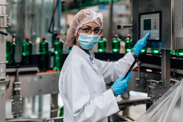 Beautiful female worker in workwear, with disposable protective cap and with protective mask on her face working in bottling factory. Inspection quality control.