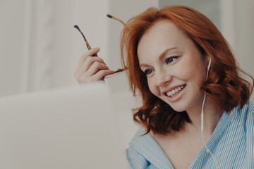Close up shot of redhead businesswoman makes video call to business partner, has discussion with corporate partner, holds optical glasses, smiles pleasantly, uses earphones. Remote job interview
