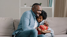Happy Little Daughter Sitting On Sofa With Closed Eyes Receiving Gift Box From Loving Father Caring Dad Congratulating Kid Girl Making Birthday Present Surprise Daddy Hugging Cute Child Cuddling Baby