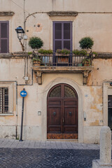 Bottom-up view to balcony with metal designed railing and with flower pots on it, with black outdoor lantern and electrical wires on ancient building wall, and with window shutters in background