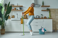 Playful Senior Woman Dancing With A Mop While Standing At The Domestic Kitchen