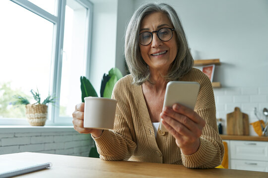Cheerful senior woman enjoying hot drink and using smart phone while relaxing at home