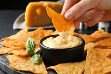 Woman Dipping Crispy Nacho Into Delicious Cheese Sauce At Table, Closeup