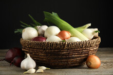 Wicker Basket With Fresh Onion Bulbs, Leeks And Garlic On Wooden Table