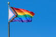 © madscinbca - A LGBTQ+ rainbow progress pride flag flaps from a flagpole in a brisk breeze. Blue sky behind.