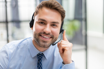 Sticker - Cheerful young support phone male operator in headset, at workplace with collegues on the background while using computer, help service and client consulting call center concept.