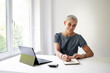 © epiximages - pretty middle aged woman with grey dress sits in white office by desk with short hair and works on laptop