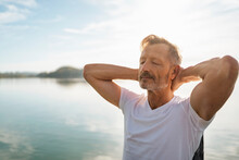 Mature Man With Eyes Closed And Hands Behind Head By Lake On Sunny Day