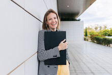 Happy Businesswoman Holding File Folder Leaning On Wall