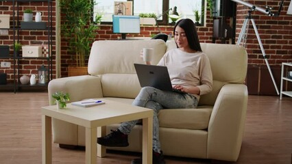 Poster - Focused young adult asian student attending online class while typing important lessons on modern portable computer at home. Beautiful woman sitting on sofa while working remotely on laptop.