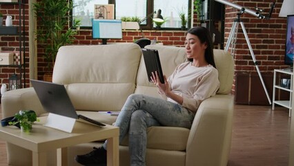 Poster - Happy asian woman having modern tablet browsing internet while sitting on couch at home. Cheerful smiling heartily person with touchscreen device surfing webpages while doing remote work.