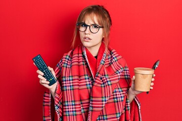 Wall Mural - Redhead young woman wrapped in a red warm red blanket eating icecream and watching tv clueless and confused expression. doubt concept.