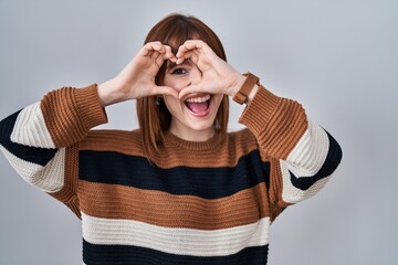 Sticker - Young beautiful woman wearing striped sweater over isolated background doing heart shape with hand and fingers smiling looking through sign