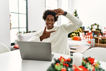 Poster - Young african american man using laptop sitting on the table by christmas tree smiling making frame with hands and fingers with happy face. creativity and photography concept.