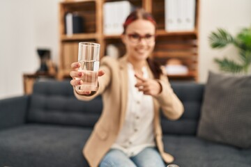 Canvas Print - Young caucasian woman offering a glass of water smiling happy pointing with hand and finger