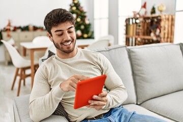 Sticker - Young hispanic man smiling happy sitting on the sofa using touchpad at home.