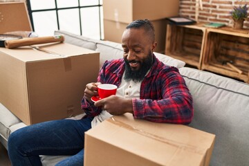 Poster - Young african american man drinking coffee sitting on sofa at new home