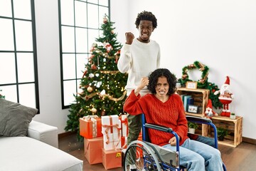 Poster - Young interracial couple with woman sitting on wheelchair by christmas tree angry and mad raising fist frustrated and furious while shouting with anger. rage and aggressive concept.