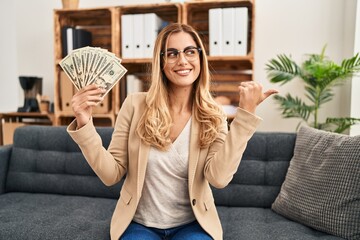 Poster - Young blonde therapist woman working at therapy office holding money pointing thumb up to the side smiling happy with open mouth