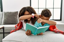 Two Siblings Lying On The Sofa Reading A Book Smiling Cheerful Playing Peek A Boo With Hands Showing Face. Surprised And Exited