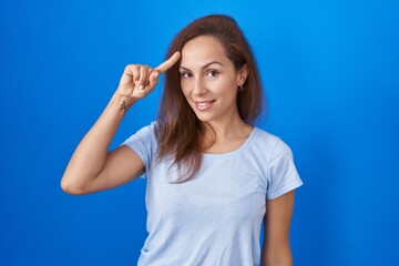 Canvas Print - Brunette woman standing over blue background smiling pointing to head with one finger, great idea or thought, good memory