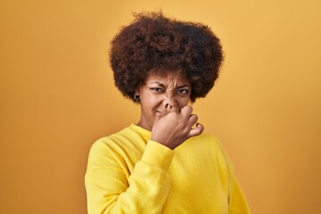 Poster - Young african american woman standing over yellow background smelling something stinky and disgusting, intolerable smell, holding breath with fingers on nose. bad smell