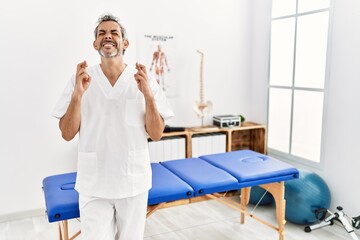 Sticker - Middle age hispanic therapist man working at pain recovery clinic gesturing finger crossed smiling with hope and eyes closed. luck and superstitious concept.