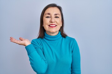 Sticker - Middle age hispanic woman standing over isolated background smiling cheerful presenting and pointing with palm of hand looking at the camera.