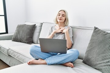 Sticker - Young caucasian woman using laptop at home sitting on the sofa smiling with hands on chest with closed eyes and grateful gesture on face. health concept.