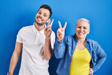 Sticker - Young brazilian mother and son standing over blue background smiling looking to the camera showing fingers doing victory sign. number two.