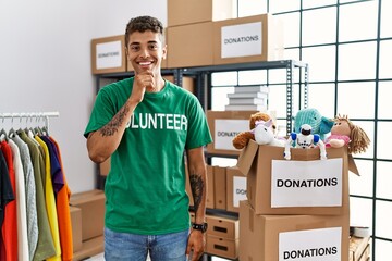 Canvas Print - Young handsome hispanic man wearing volunteer t shirt at donations stand with hand on chin thinking about question, pensive expression. smiling and thoughtful face. doubt concept.