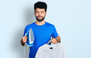 Poster - Young arab man with beard holding electric steam iron and white t shirt smiling looking to the side and staring away thinking.