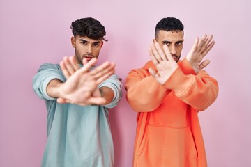 Sticker - Young hispanic gay couple standing over pink background rejection expression crossing arms and palms doing negative sign, angry face